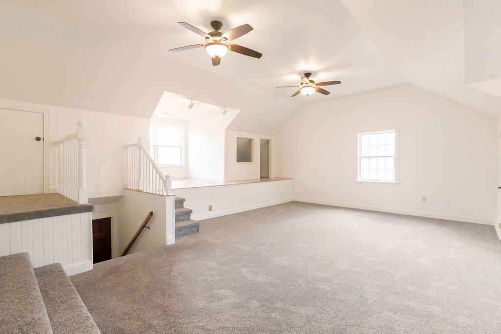 Spacious loft area featuring vaulted ceilings, two ceiling fans with integrated lighting, a dedicated elevated platform with track lighting and windows, and carpeting throughout