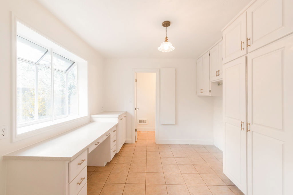 Bright room featuring a large bay window, extensive built-in desk space, white cabinetry with brass hardware, and tile flooring