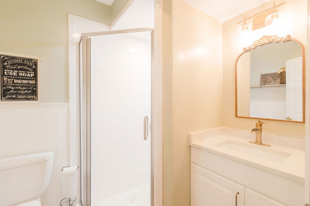 Bathroom featuring a shower enclosure with a clear glass door, white wainscoting, a vanity with a white countertop and integrated sink, a gold-toned faucet, and an ornate framed mirror