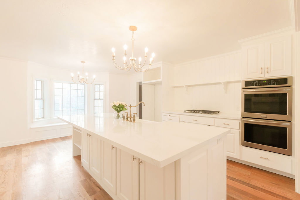 Spacious kitchen featuring a large central island with an integrated sink, double wall ovens, built-in gas cooktop, white cabinetry, and wood-finish flooring