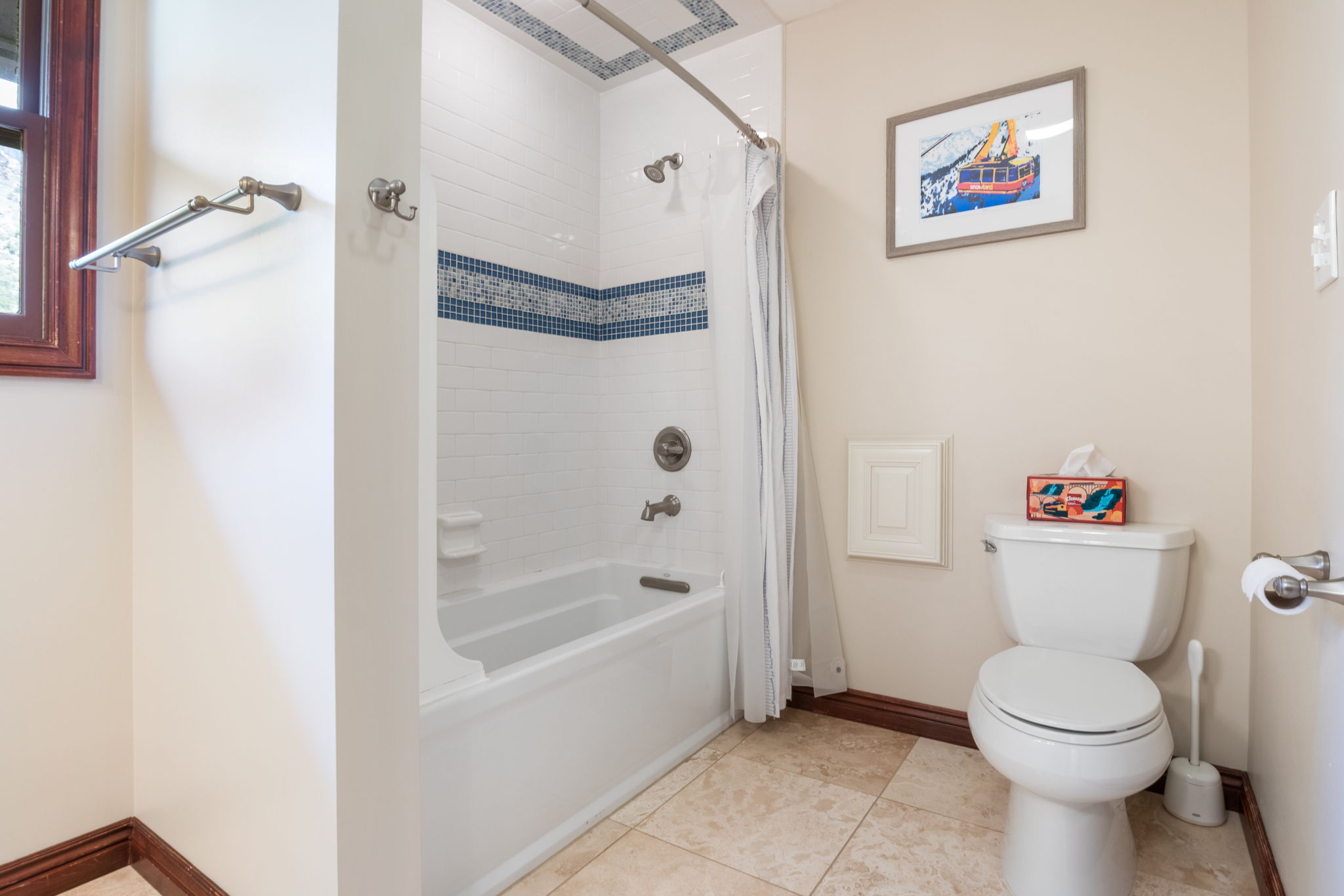 Bathroom featuring a white bathtub with tile surround, decorative blue accent tile, a window with dark wood trim, a toilet, and light-toned floor tiles