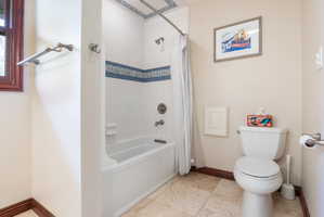Bathroom featuring a white bathtub with tile surround, decorative blue accent tile, a window with dark wood trim, a toilet, and light-toned floor tiles