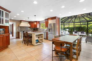 Spacious kitchen featuring a large central island with integrated shelving, tile flooring, rich wood cabinetry, stainless steel appliances, and an attached sunroom with a glass ceiling and walls