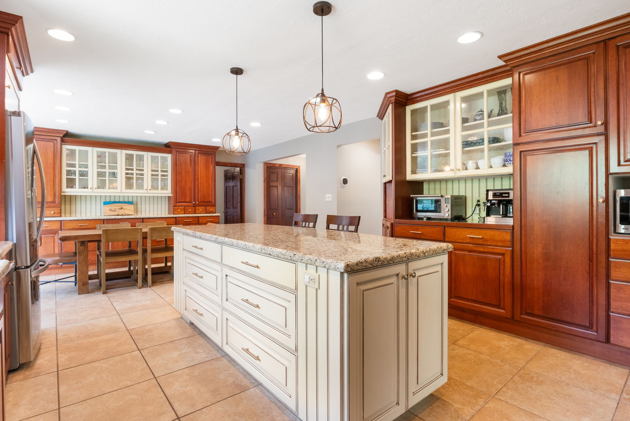 Spacious kitchen featuring a large central island with a light-colored countertop, ceramic tile flooring, extensive wood cabinetry, recessed lighting, and decorative pendant light fixtures