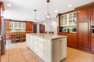 Spacious kitchen featuring a large central island with a light-colored countertop, ceramic tile flooring, extensive wood cabinetry, recessed lighting, and decorative pendant light fixtures