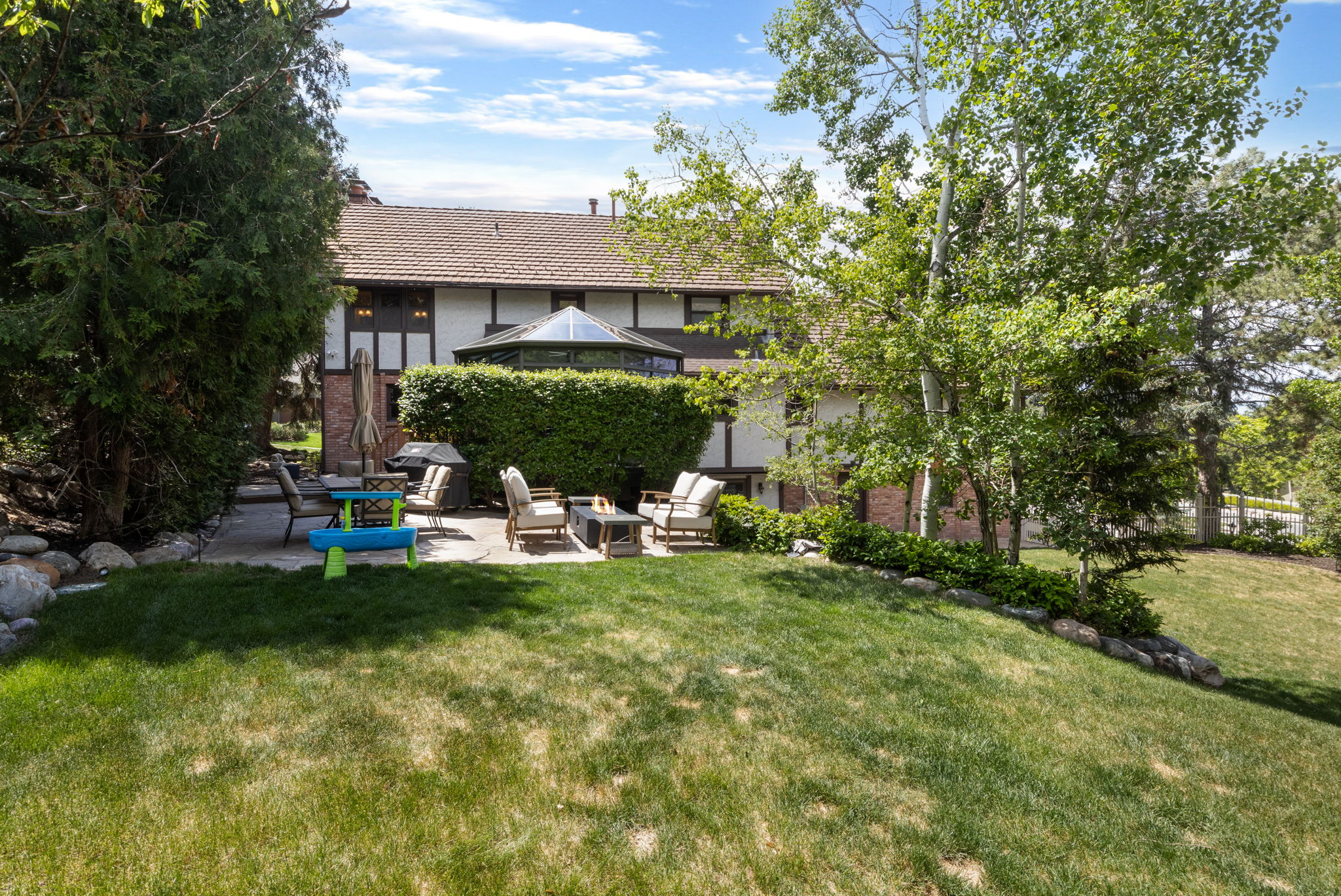 Spacious backyard featuring a manicured lawn, stone retaining walls, mature trees, and a paved patio area
