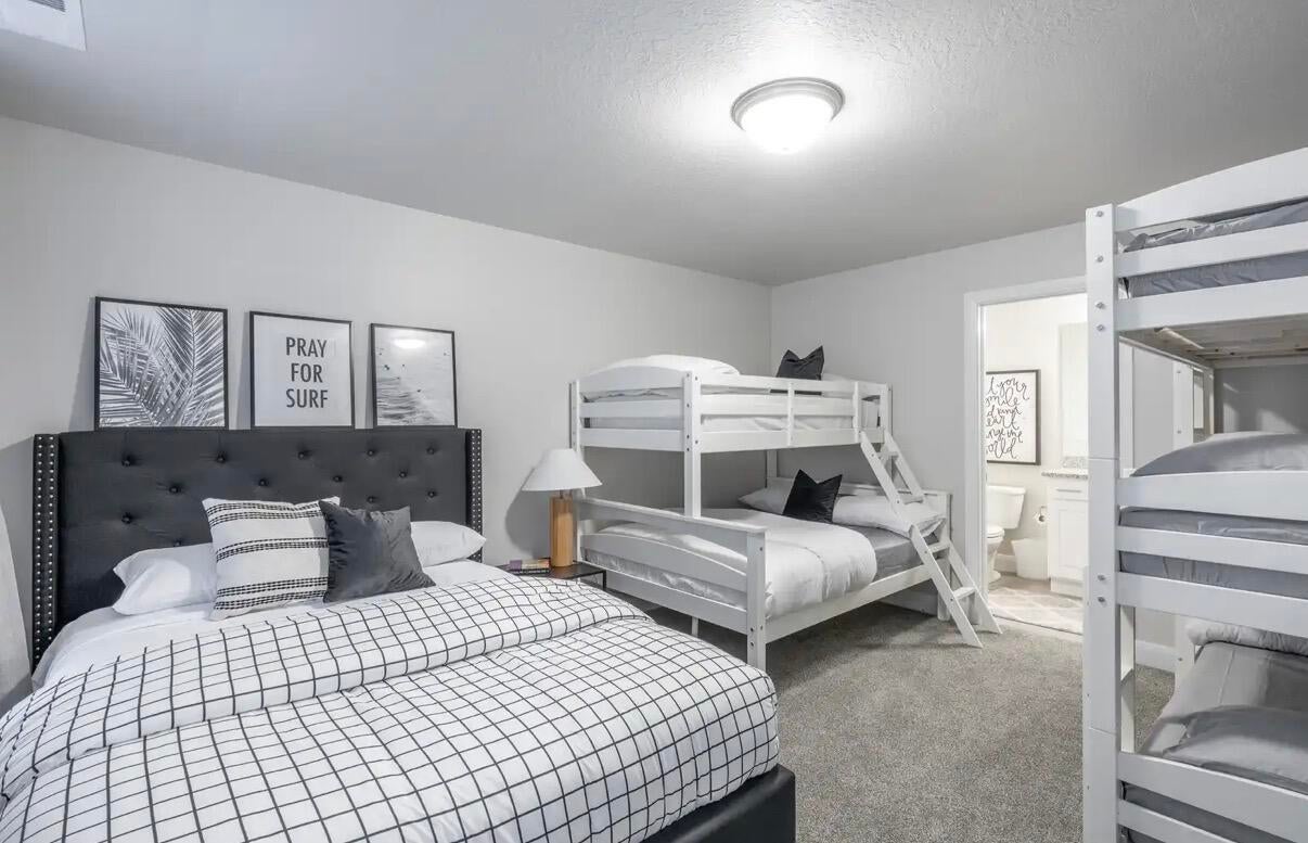 Spacious bedroom featuring neutral wall tones, plush carpet flooring, a flush-mount ceiling light, and access to a full bathroom with a vanity and toilet