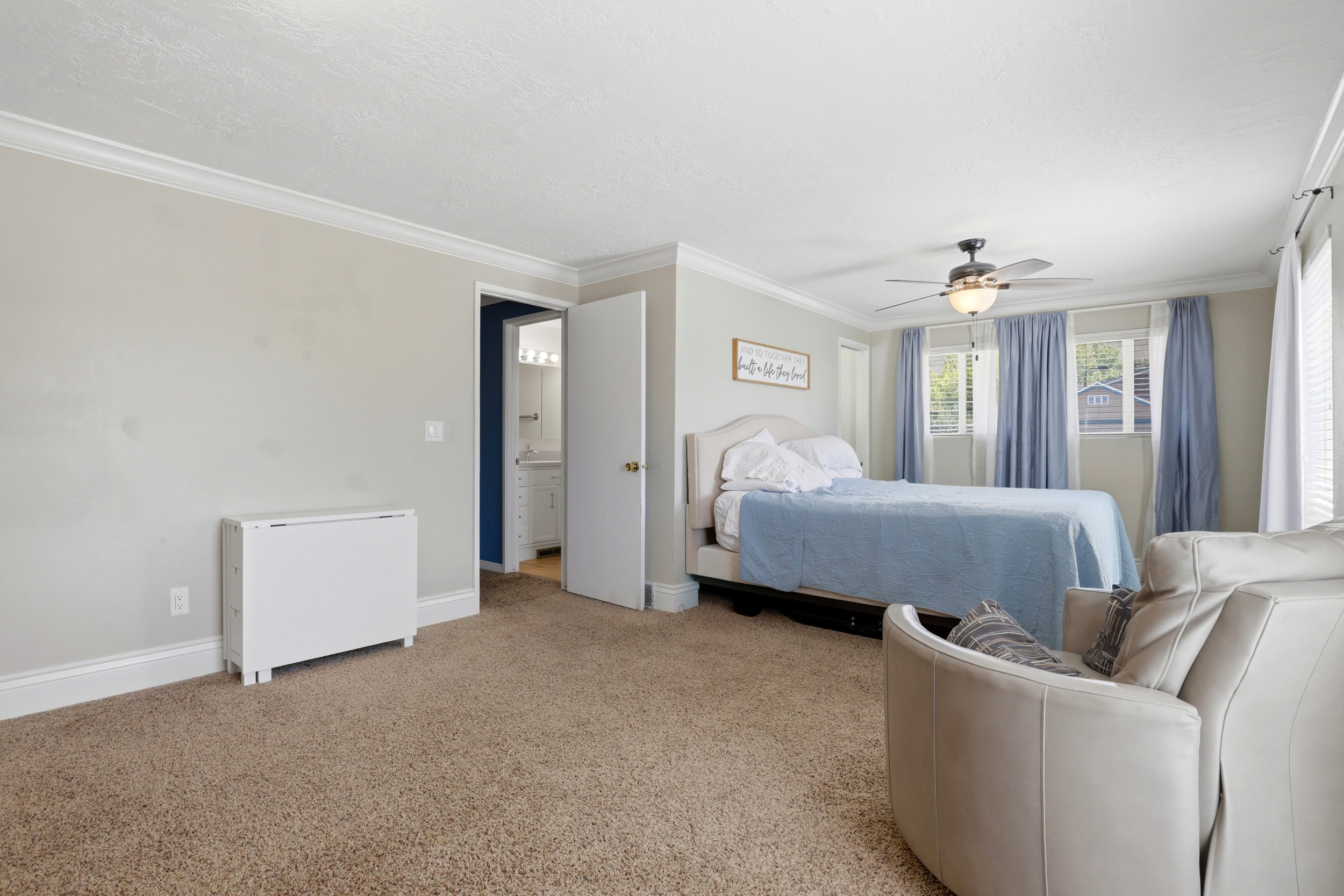 Carpeted room featuring a ceiling fan with light fixture, multiple windows, crown molding, and a visible doorway revealing a bathroom vanity with a mirror and lighting
