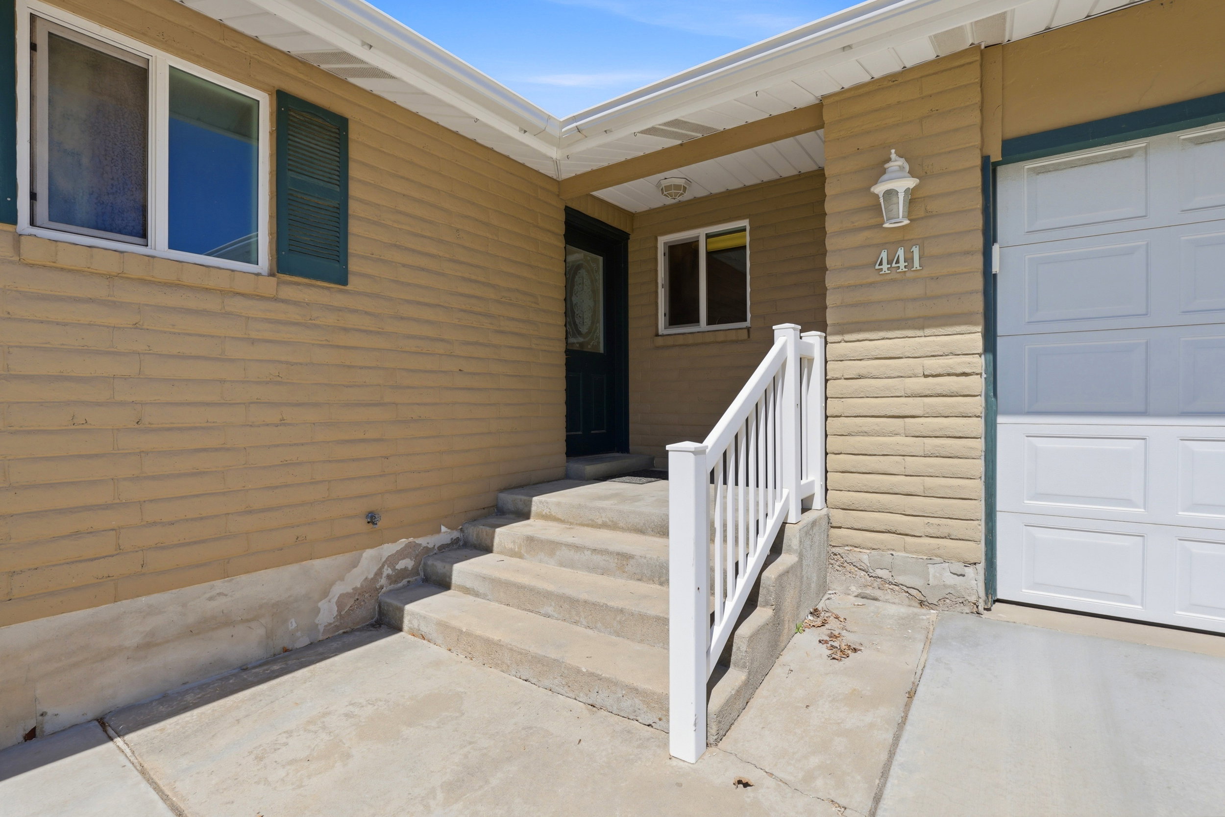 Front exterior featuring tan brick siding, white window trim, dark green shutters, a white garage door, and concrete steps with a white railing