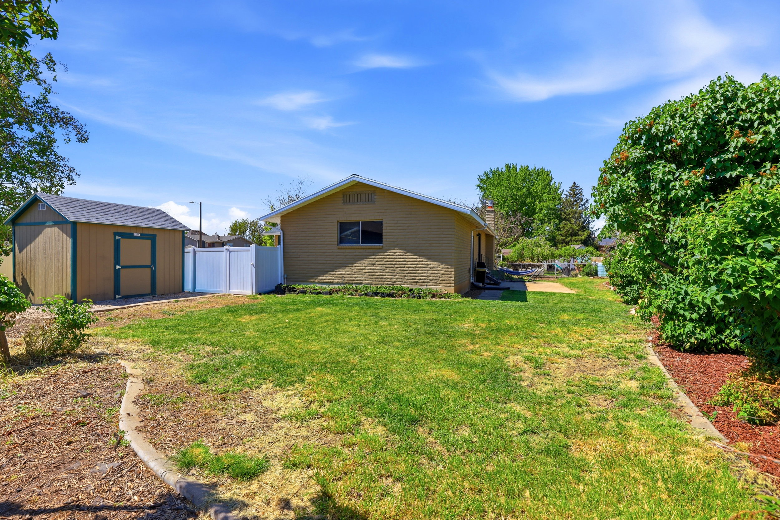 Expansive backyard featuring a green lawn, a detached storage shed with a shingle roof, a white vinyl fence, and mature perimeter landscaping