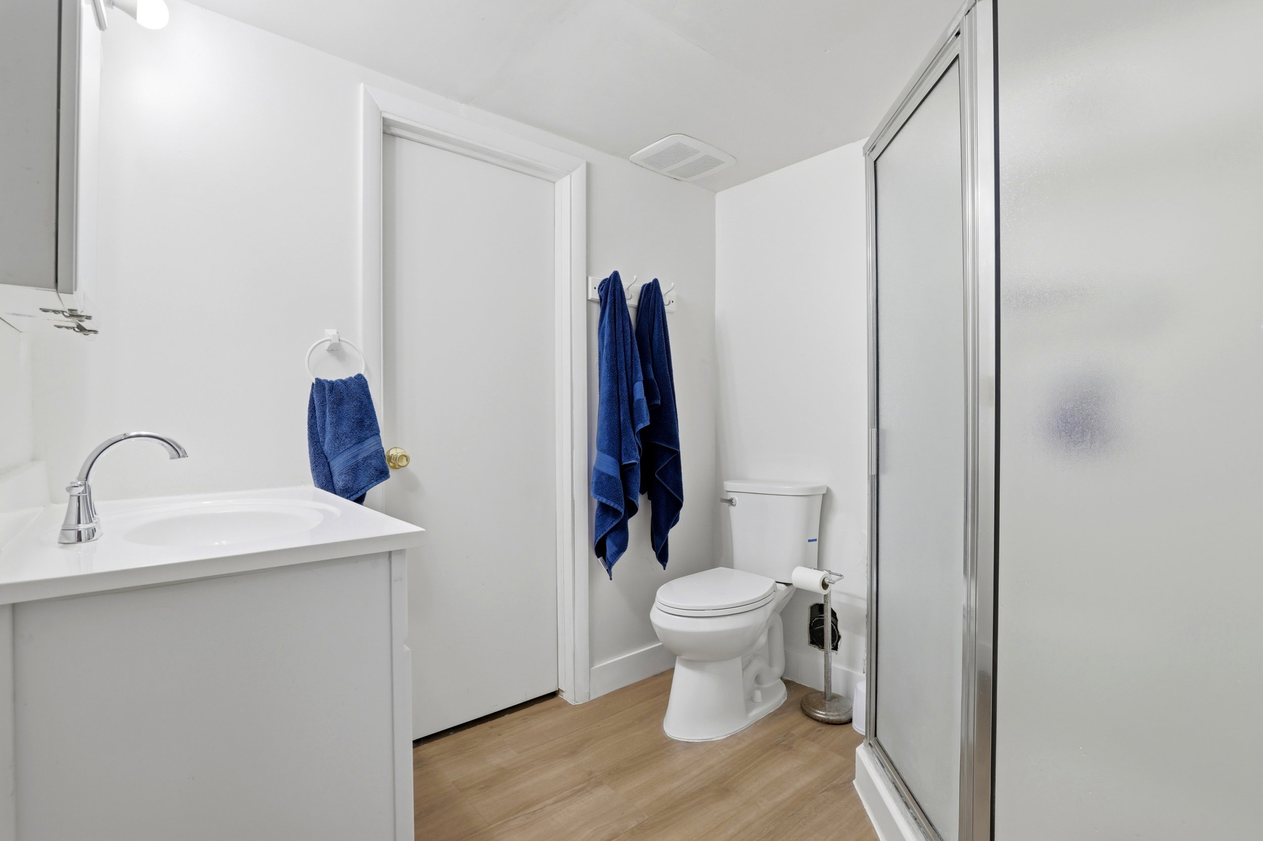 Bathroom featuring a single vanity with a white countertop and chrome faucet, wood-finish flooring, a toilet, and a stand-up shower with a frosted glass door