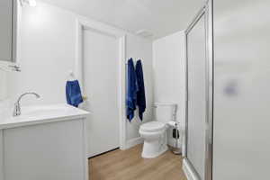 Bathroom featuring a single vanity with a white countertop and chrome faucet, wood-finish flooring, a toilet, and a stand-up shower with a frosted glass door