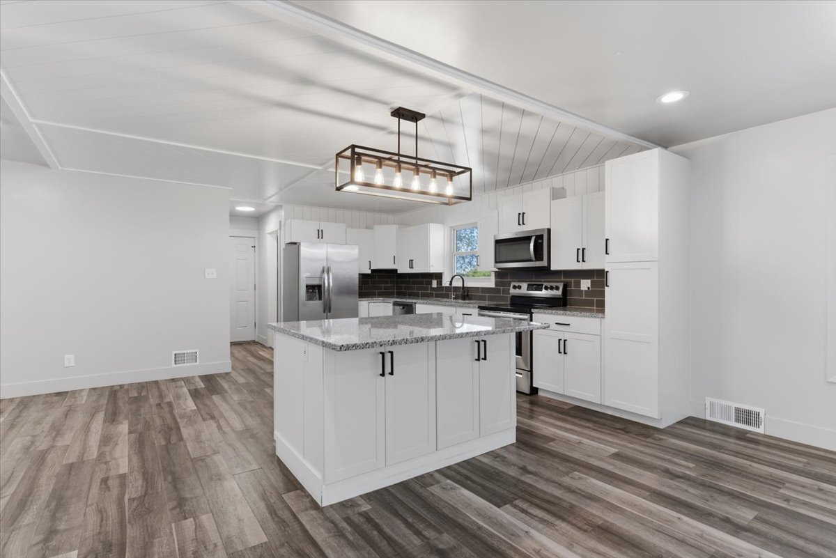 Open-concept kitchen featuring a large island with granite countertops, vinyl flooring, white cabinetry with black hardware, included stainless steel appliances, and a dark tile backsplash