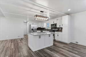 Open-concept kitchen featuring a large island with granite countertops, vinyl flooring, white cabinetry with black hardware, included stainless steel appliances, and a dark tile backsplash