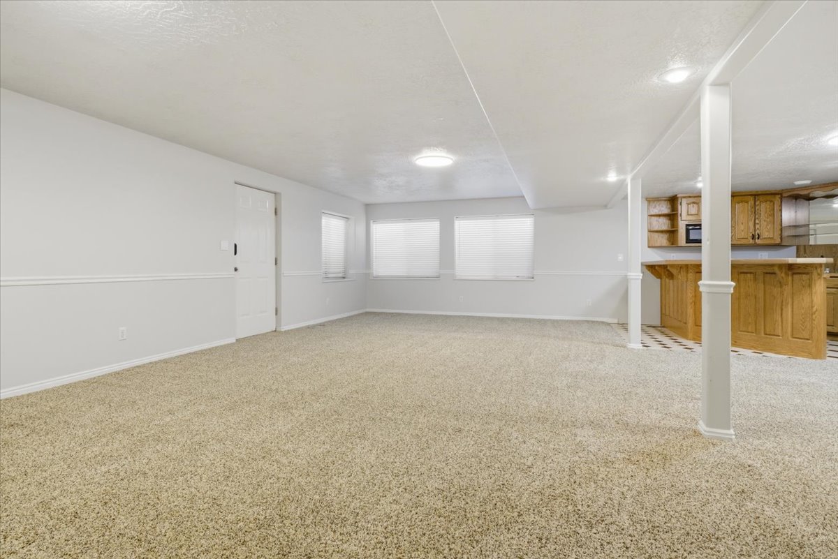 Spacious carpeted room featuring a white door, two windows with blinds, a built-in wet bar with wooden cabinetry, and recessed lighting