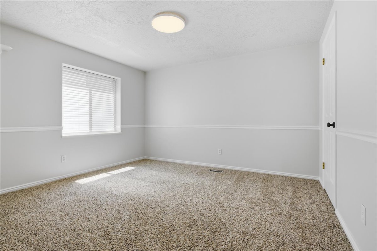 Carpeted room featuring a window with blinds, white walls, chair rail molding, a white paneled door with a black doorknob, and a flush mount ceiling light fixture
