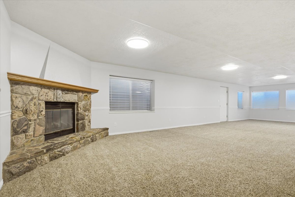 Spacious room featuring a corner stone fireplace with a wood mantel, light-colored carpeting, multiple flush-mount ceiling lights, and several windows with blinds