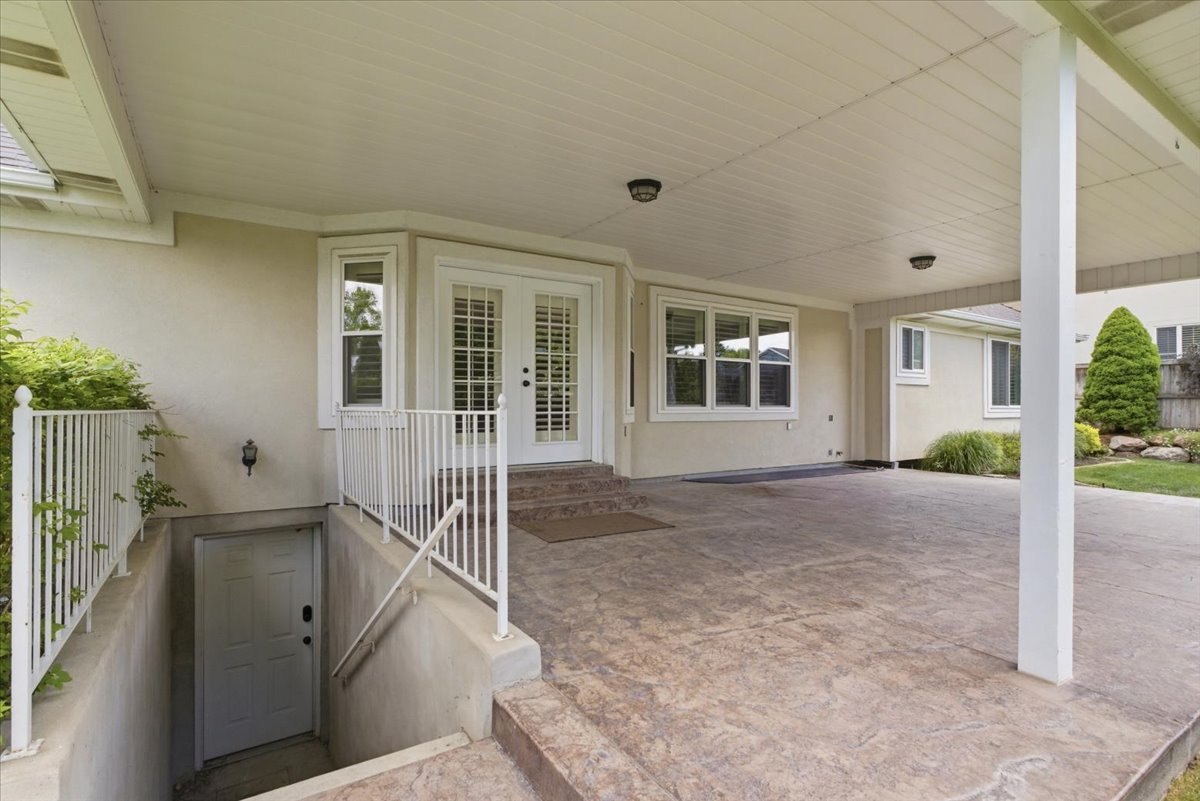 Covered patio featuring stamped concrete flooring, stucco exterior, multiple windows, and a separate basement entrance with white railings