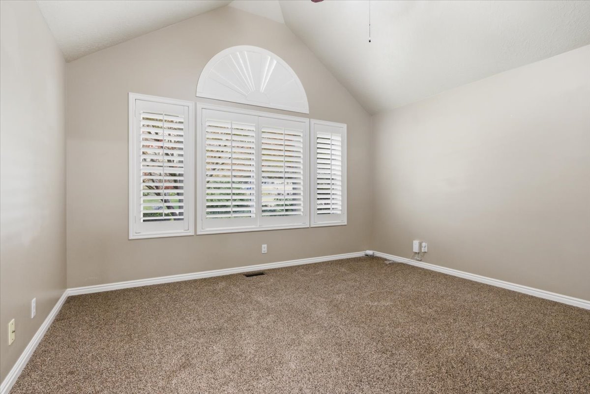 Vaulted ceiling room featuring a large window with plantation shutters, an arched transom window, and neutral carpeting