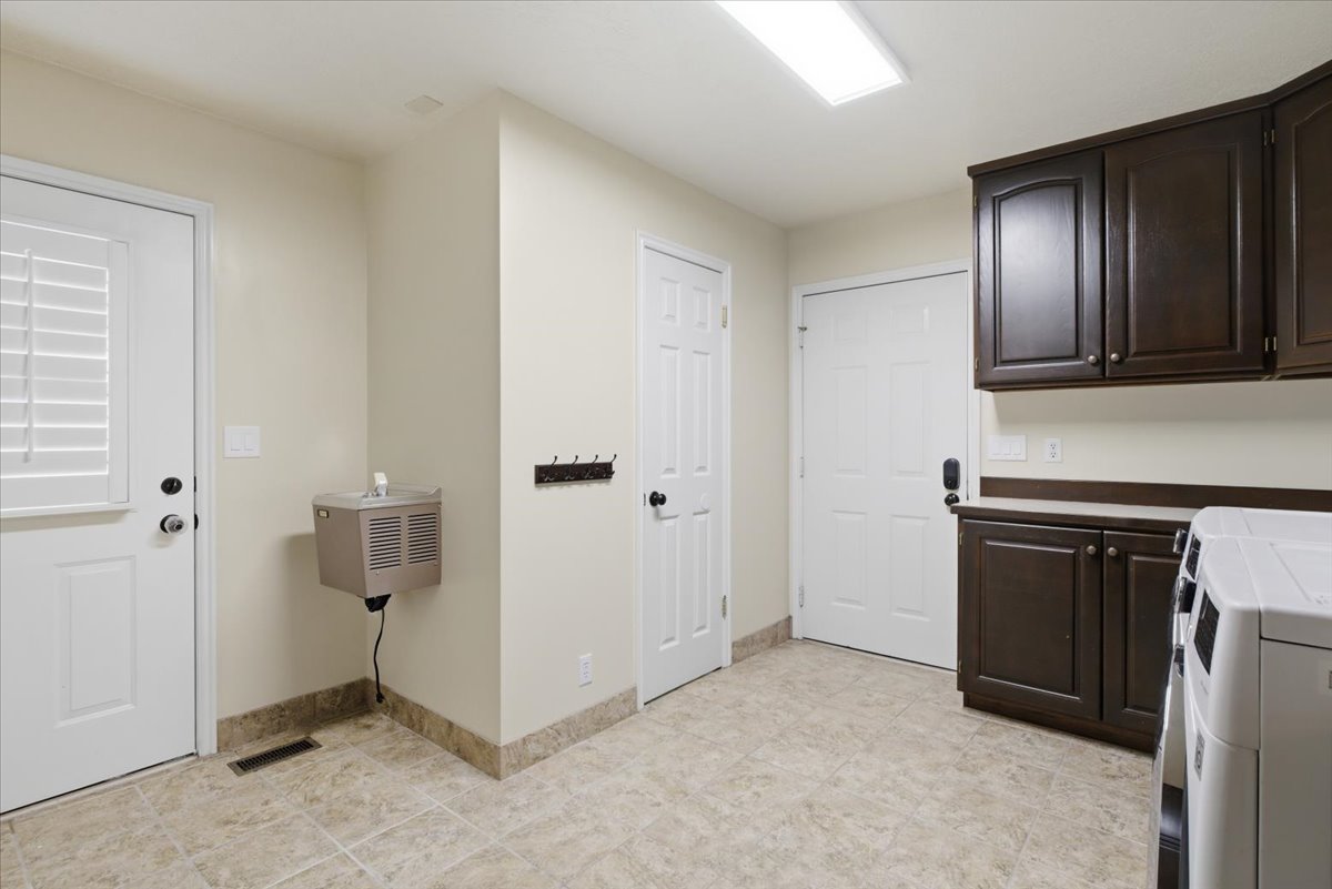 Spacious utility area featuring ceramic tile flooring, dark wood-finish cabinetry, light-toned countertops, and a wall-mounted water dispenser