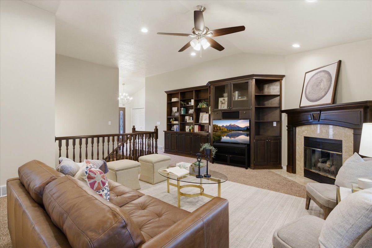 Living area featuring vaulted ceilings, a ceiling fan with integrated lighting, a built-in media center with shelving and glass-front cabinets, a fireplace with a stone surround and wood mantle, and a wood spindle railing
