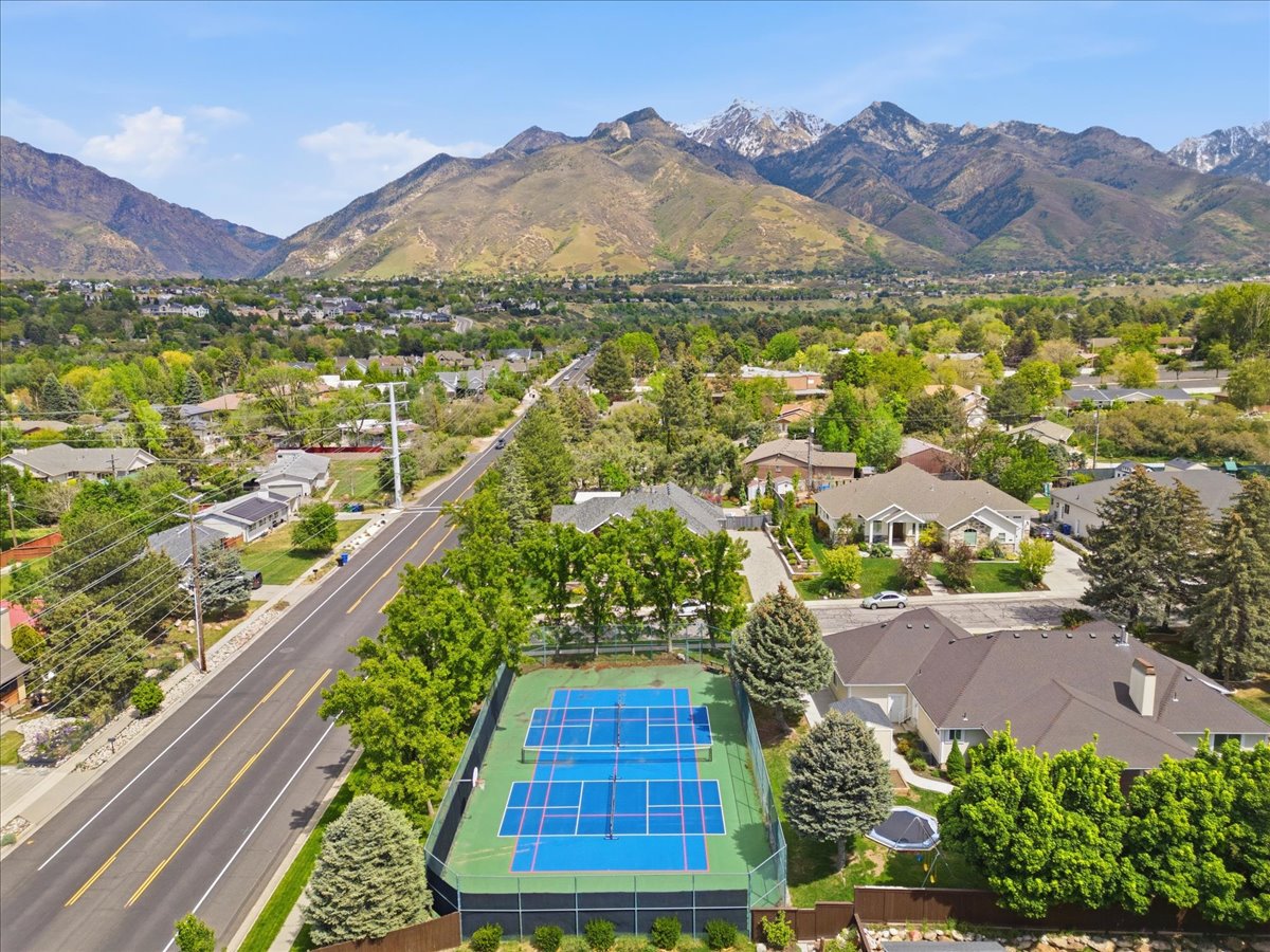 Fenced tennis court featuring two distinct playing areas with surrounding green space, situated within a residential area with mature trees and mountain scenery