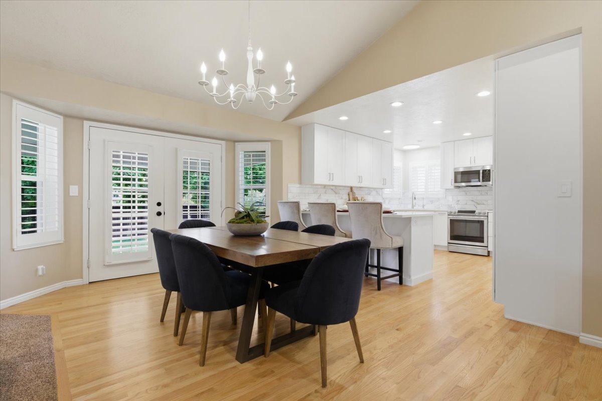Open-concept dining area featuring wood-finish flooring, a vaulted ceiling with a white chandelier, and bay windows with plantation shutters