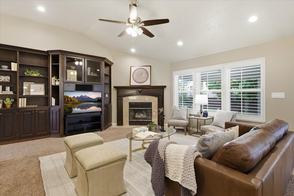 Vaulted ceiling living area featuring a dark wood built-in entertainment center, a fireplace with a decorative mantel, white plantation shutters, recessed lighting, and a ceiling fan