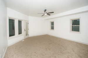 Spacious room with plush carpeting, a contemporary ceiling fan, multiple windows for natural illumination, a glass-paneled exterior door, and a tray ceiling detail