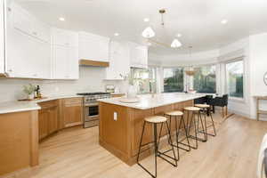 Open-concept kitchen featuring a large central island with seating, a professional-grade stainless steel range, light wood-finish flooring, two-tone cabinetry with white upper and wood-tone lower cabinets, and a bay window dining area