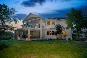 Expansive rear elevation featuring a covered patio with stone-clad columns, an upper-level balcony, a bay window, a large lawn, and mature trees