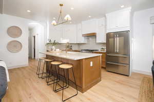 Bright kitchen featuring a large central island with seating, light-toned wood-finish flooring, white upper cabinetry, and stainless steel appliances