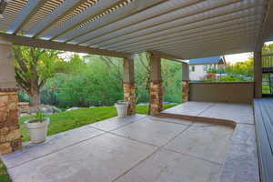 Expansive outdoor patio featuring a slatted pergola, stone-clad support columns, a concrete slab, and a composite deck extension