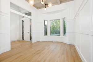 Spacious room featuring wood-finish flooring, white paneled walls, a large bay window, a vaulted ceiling with exposed beams, and a contemporary chandelier