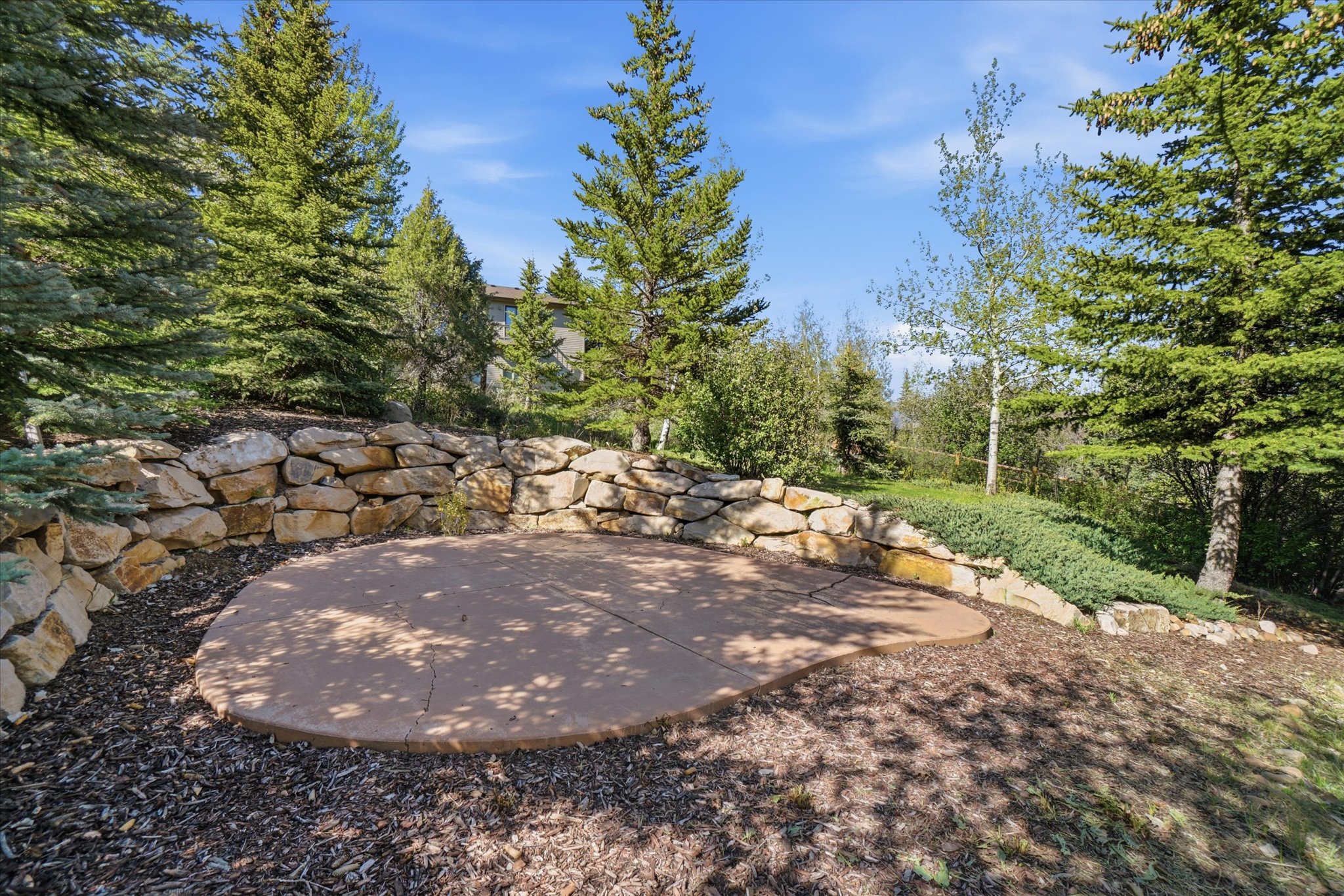 Paved outdoor patio area featuring a natural stone retaining wall, mature evergreen trees, and surrounding landscape planting