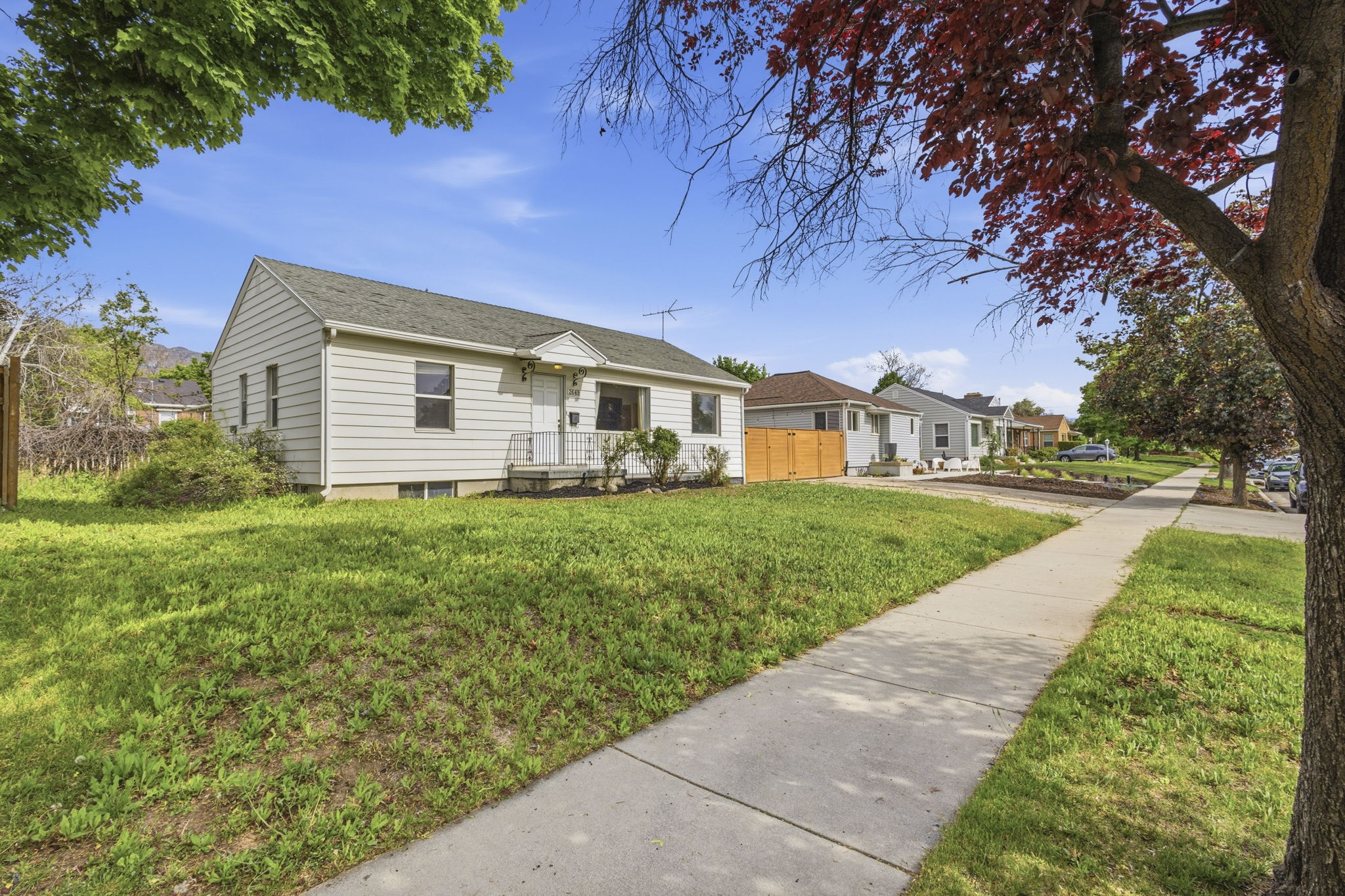 White siding exterior with a dark-pitched roof, front porch with metal railing, and a mature tree providing shade