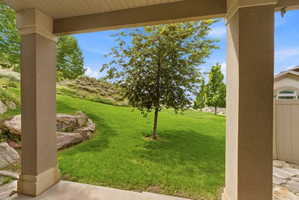 Covered patio with textured columns and a stone-look floor, overlooking a manicured lawn with mature trees and natural rock landscaping