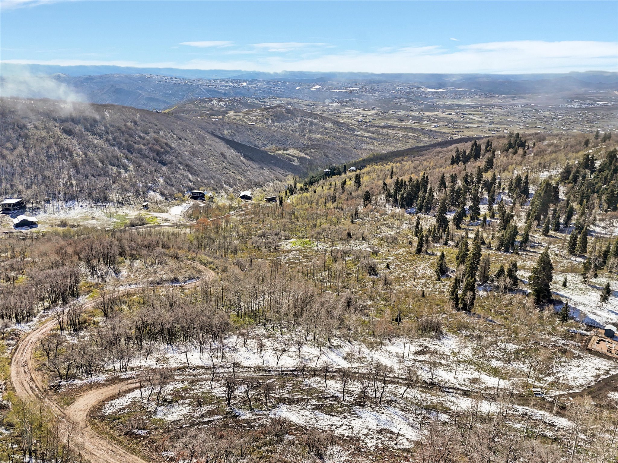 Expansive mountain landscape featuring varied terrain with a mix of deciduous and evergreen trees, a winding dirt road, and distant mountain ranges under a clear sky