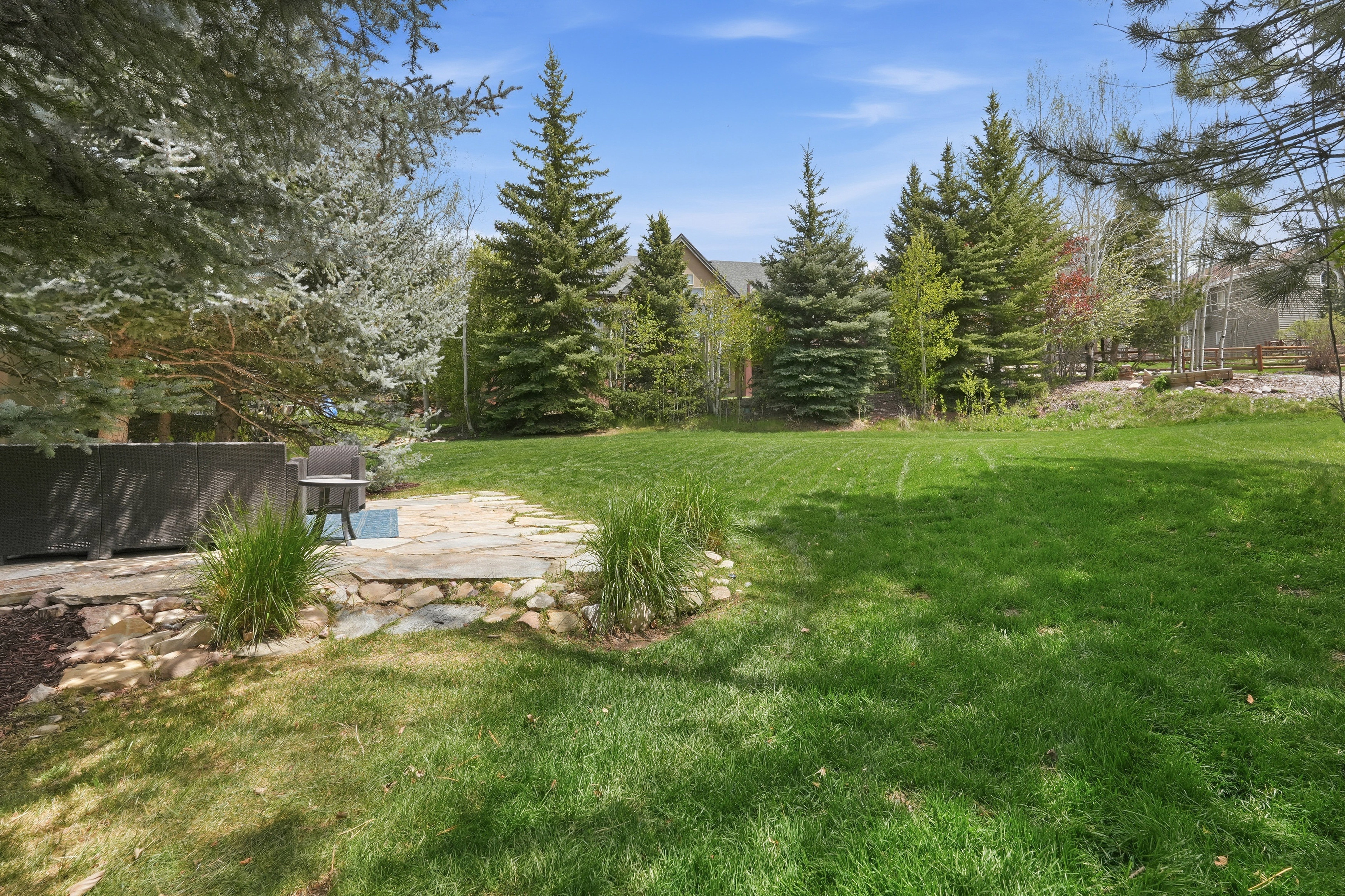 Expansive green lawn with mature evergreen trees, featuring a flagstone patio area with stone retaining wall