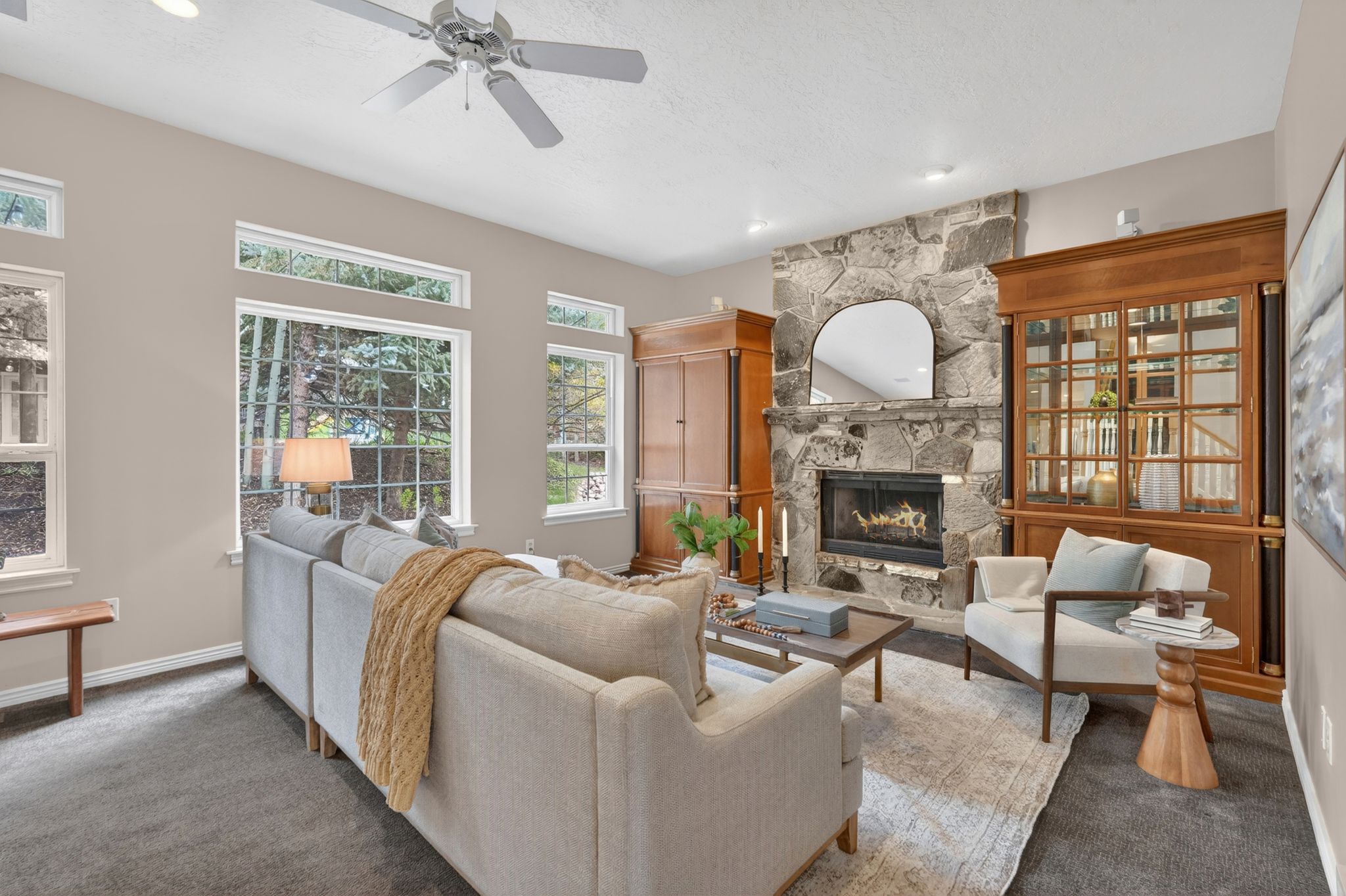 Living area featuring a stone fireplace with a wood mantel, multiple large windows, and a ceiling fan