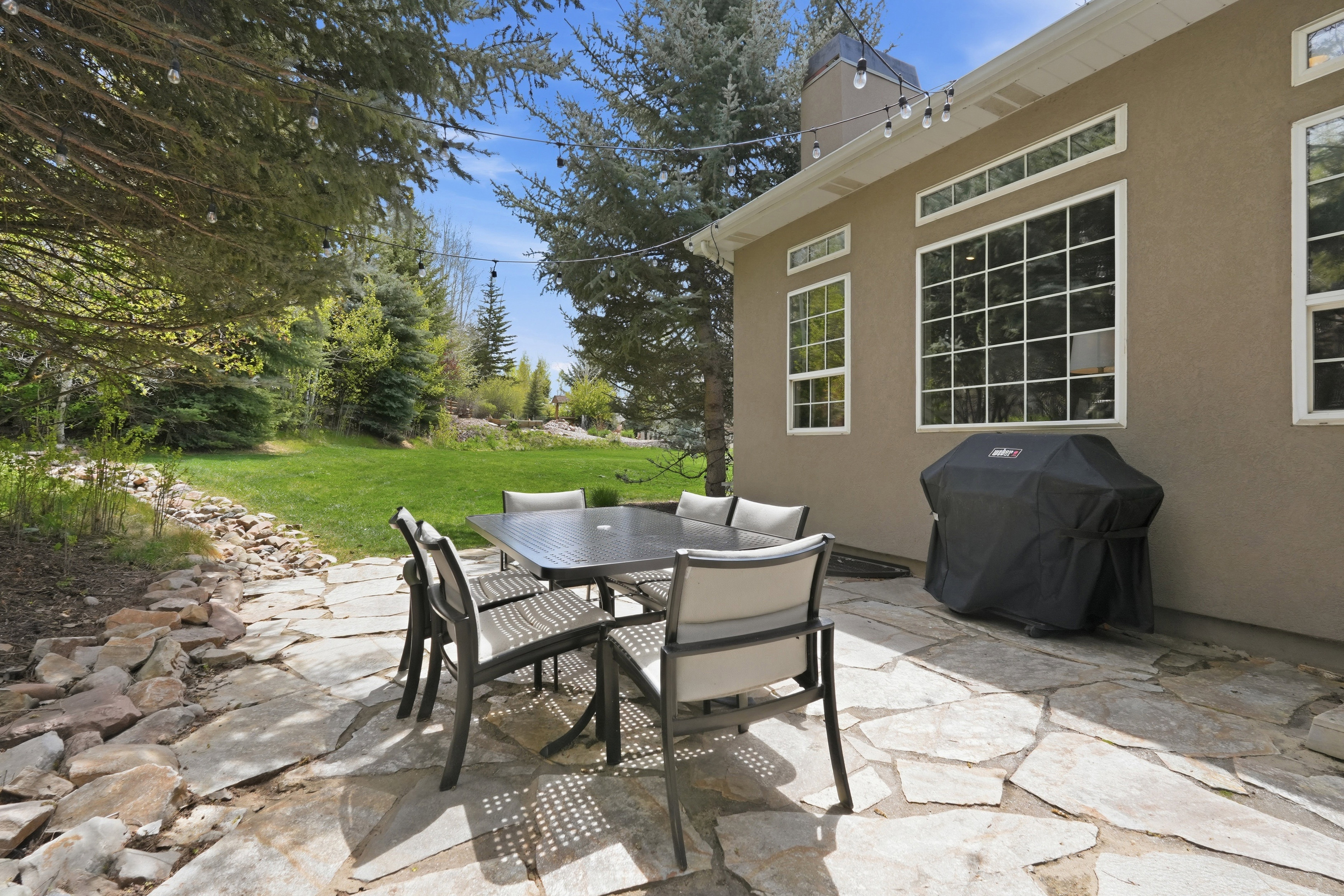 Expansive flagstone patio with a stucco exterior, multiple large windows, and a lush, landscaped yard with mature trees