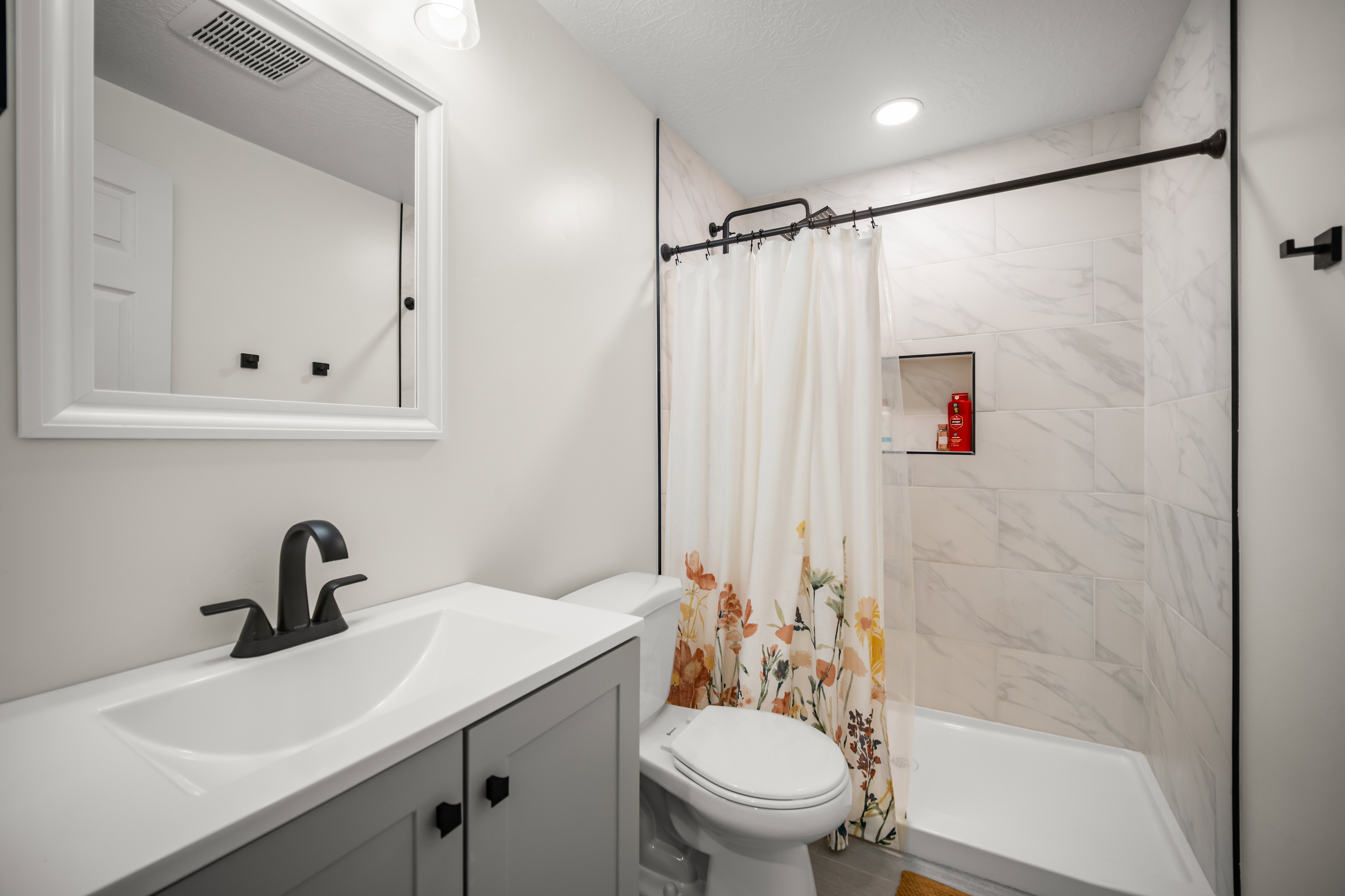 Bathroom featuring a vanity with a white countertop and dark hardware, a framed mirror, and a shower with white tile surround and a recessed niche