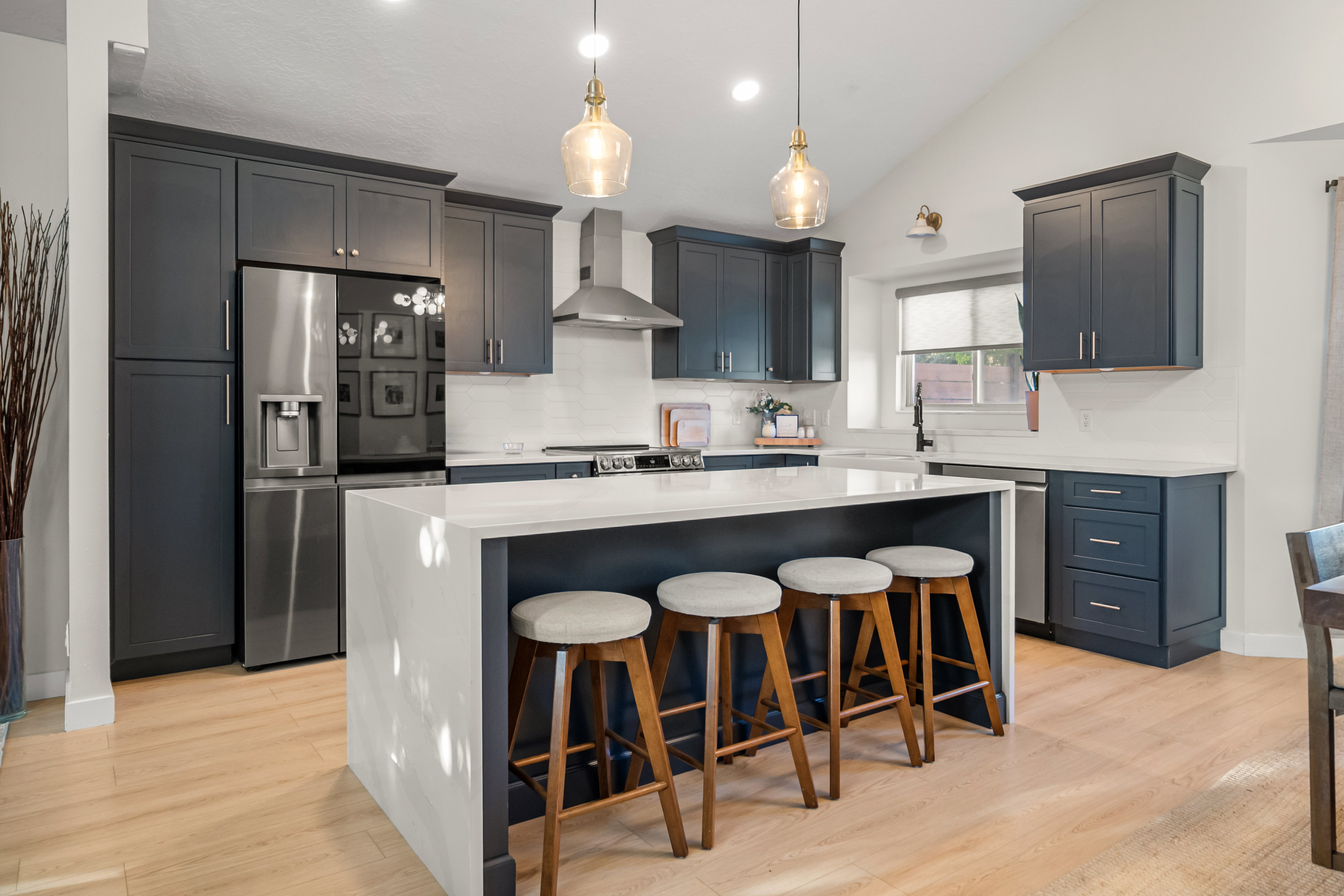 Modern kitchen featuring a waterfall island with integrated seating, dark-toned cabinetry, light wood-finish flooring, stainless steel appliances, and vaulted ceilings