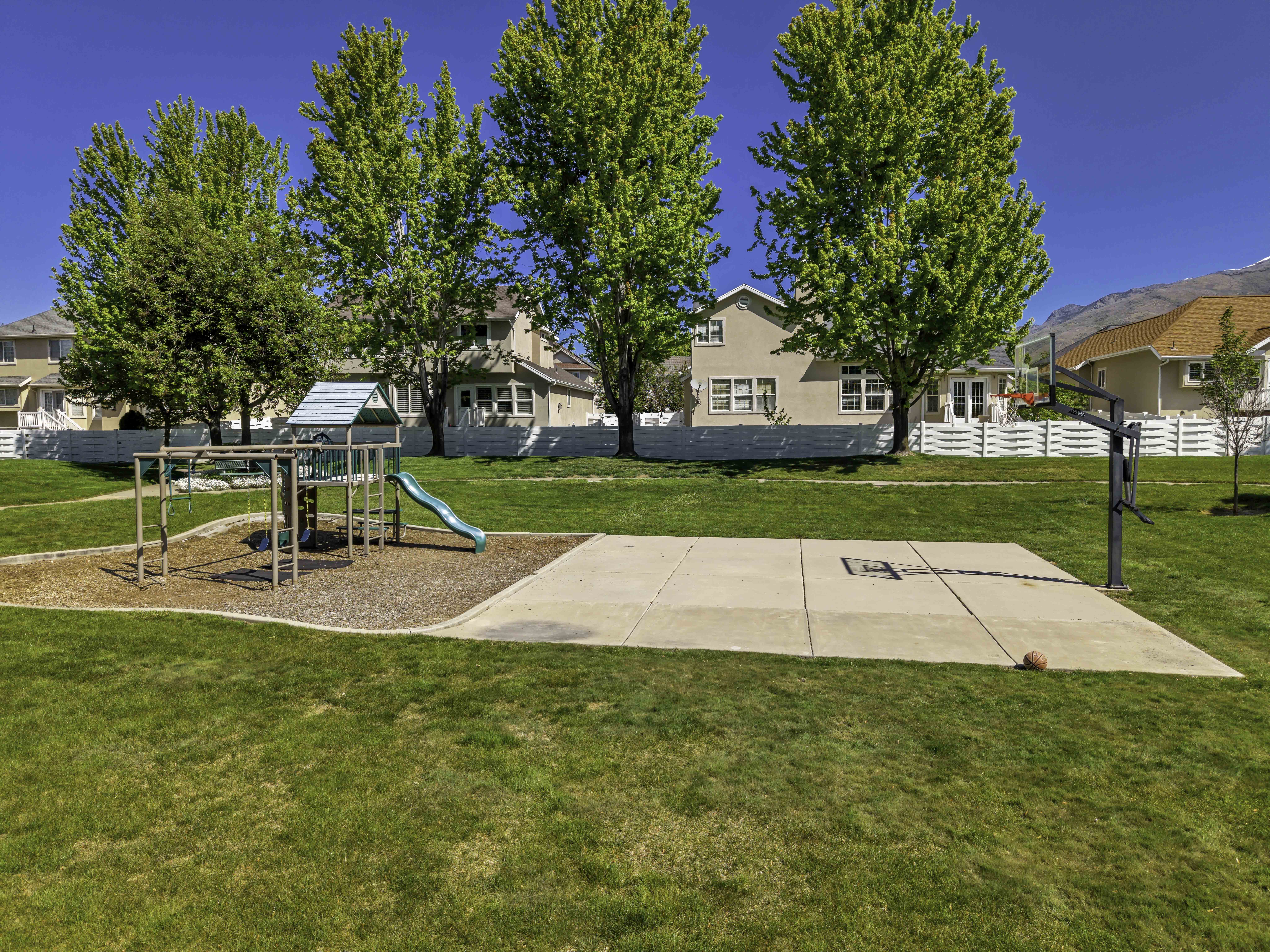 Open space featuring a playground area with a slide and swings, alongside a concrete sports court with a basketball hoop