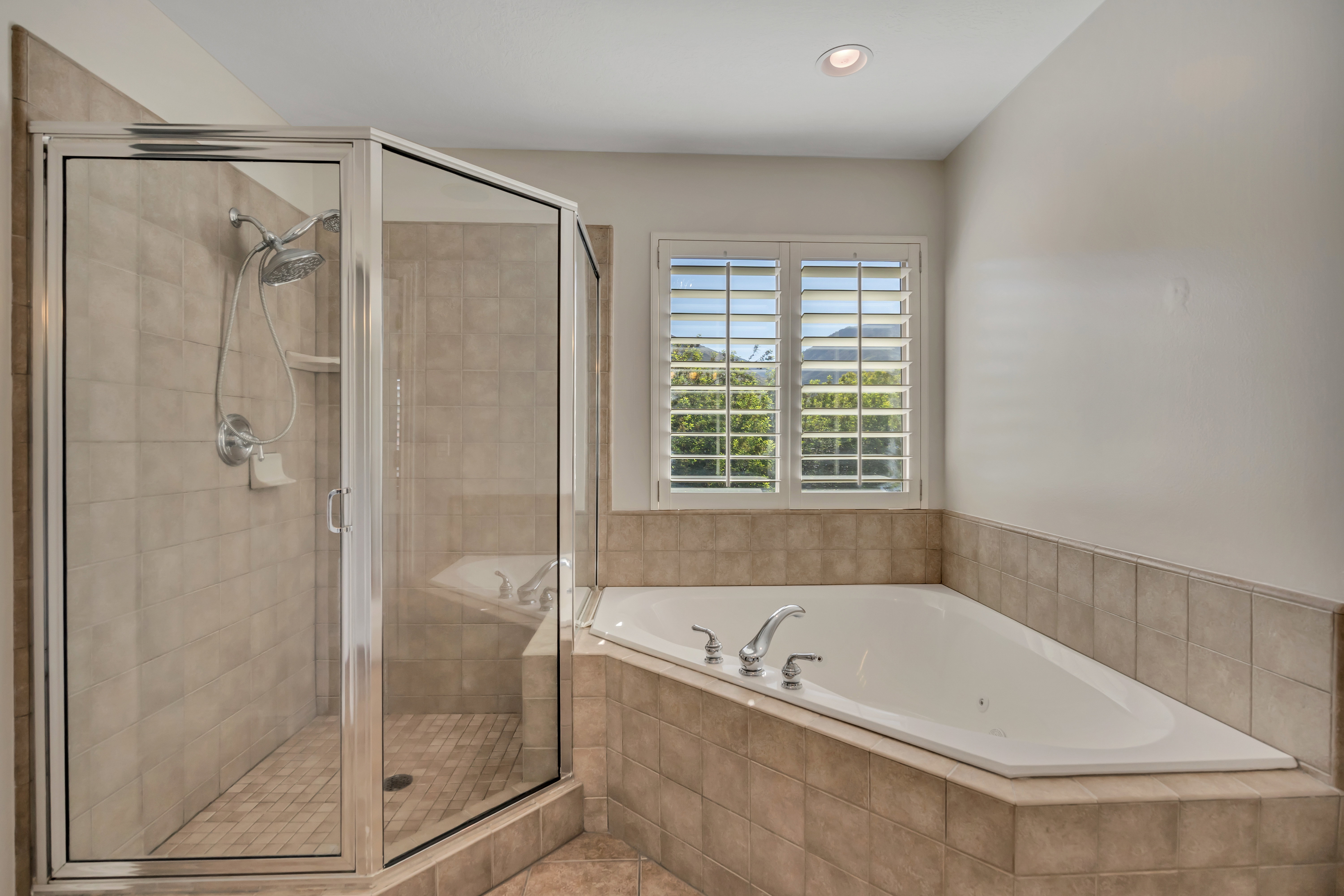 Spacious bathroom featuring a corner soaking tub with deck-mounted fixtures, a glass-enclosed shower with tile surround, and a window with plantation shutters