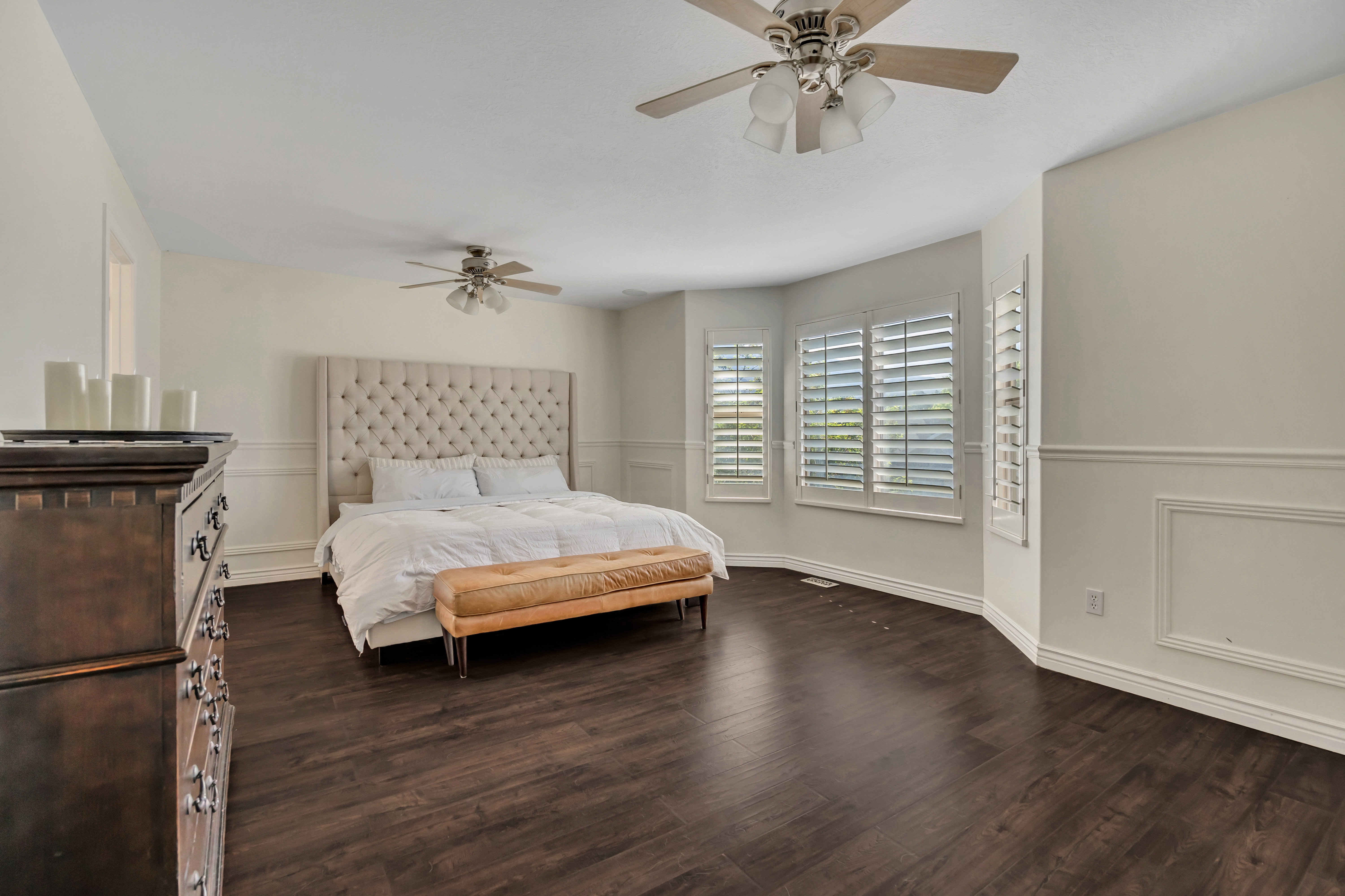 Spacious room featuring dark wood-finish flooring, multiple windows with white plantation shutters, and decorative wainscoting