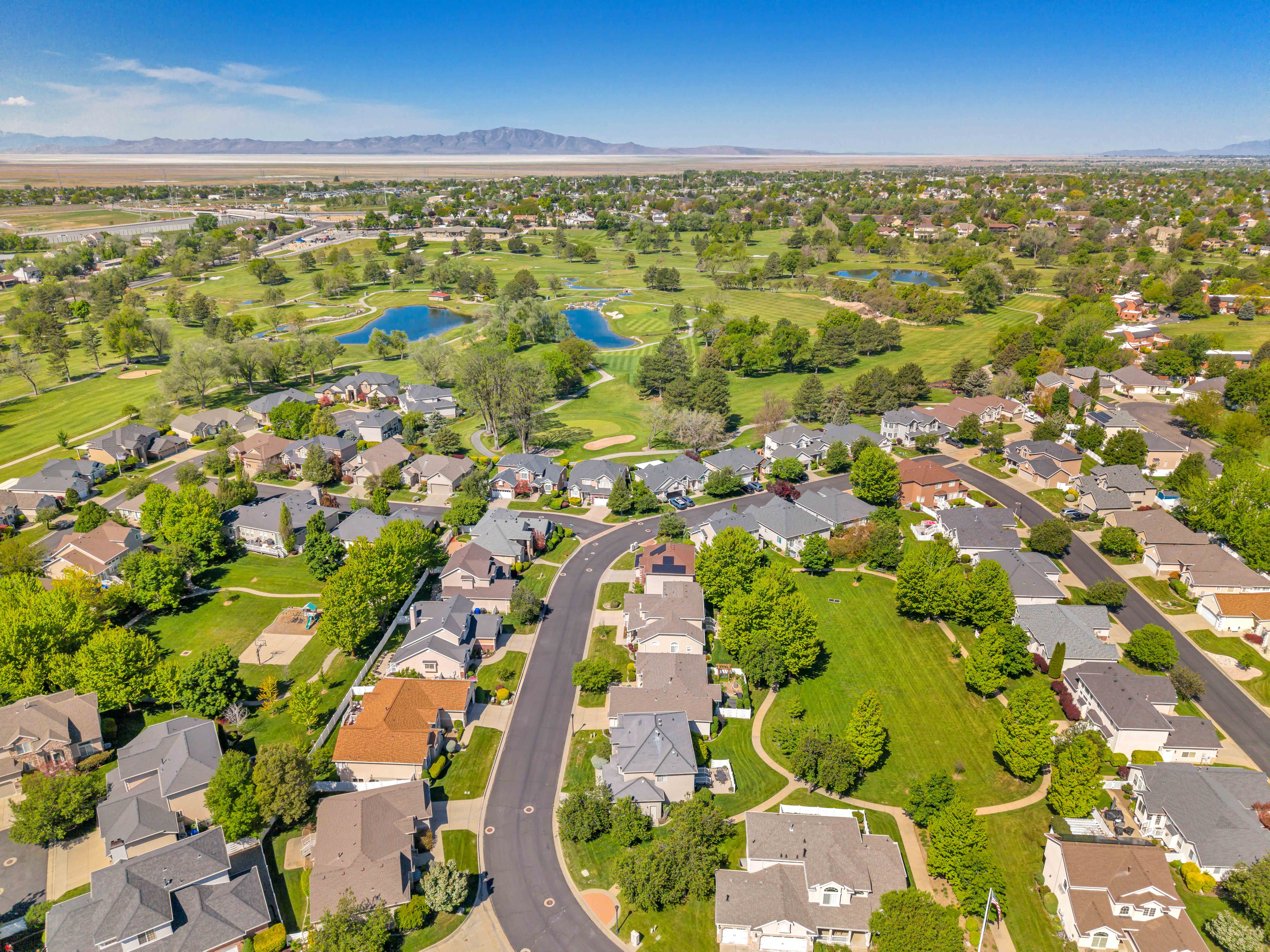 Expansive aerial panorama showcasing a residential community with varied architectural styles, mature landscaping, and an adjacent golf course featuring water features