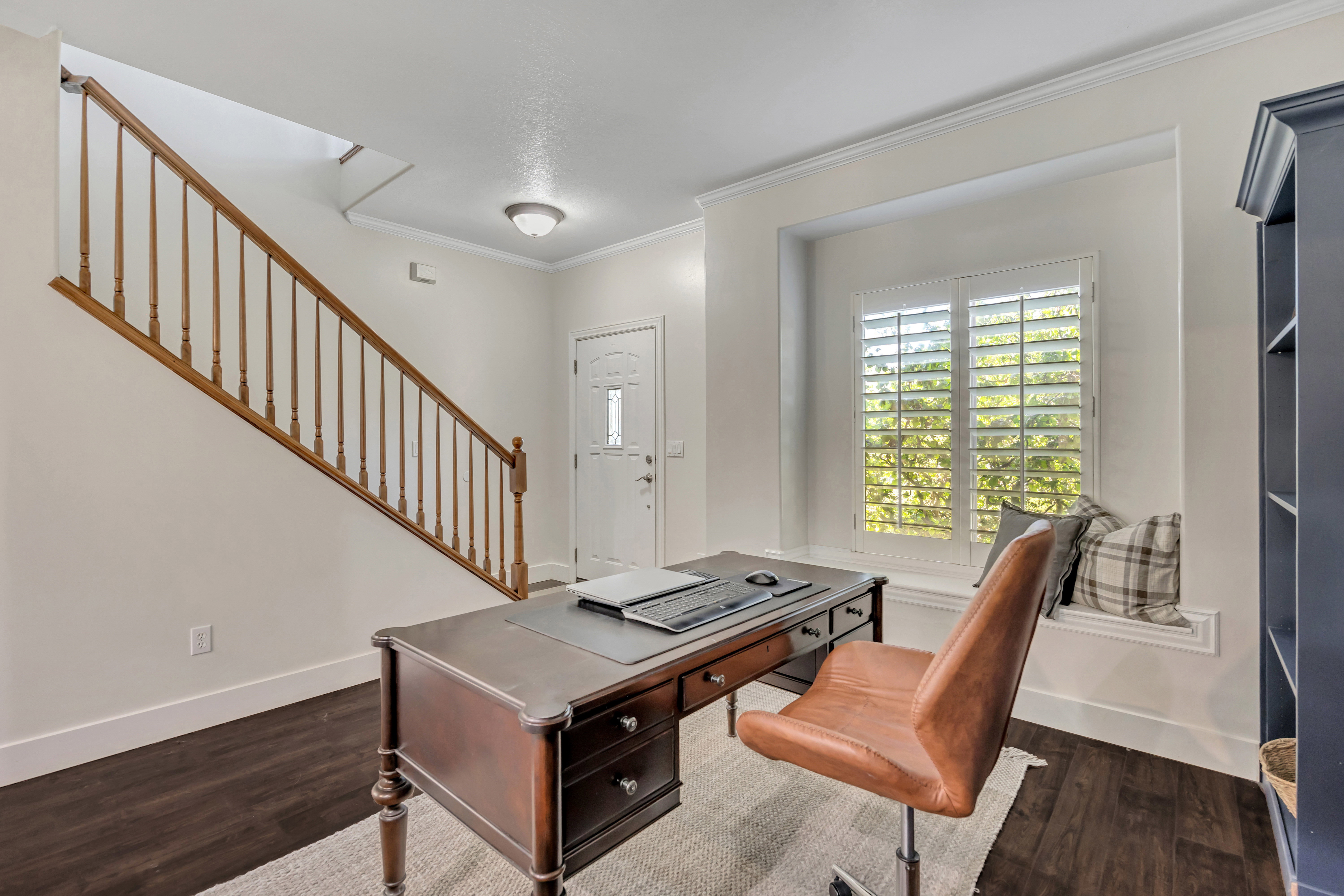 Versatile front entry area with dark wood-finish flooring, a built-in window seat, white plantation shutters, a white paneled entry door, and a staircase with a natural wood banister