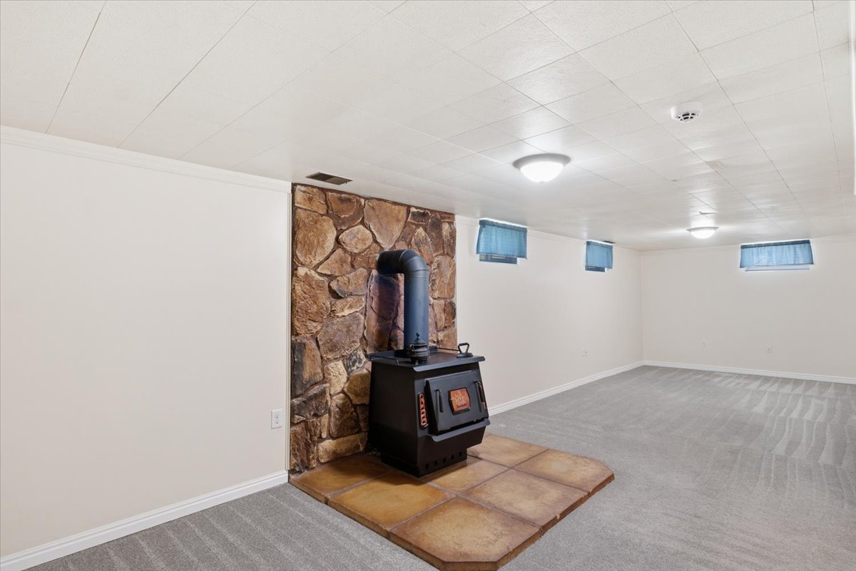 Spacious room featuring a stone accent wall with a freestanding wood-burning stove, gray patterned carpeting, white painted walls, and a white panel ceiling with flush-mount lighting fixtures