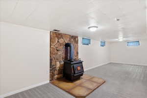 Spacious room featuring a stone accent wall with a freestanding wood-burning stove, gray patterned carpeting, white painted walls, and a white panel ceiling with flush-mount lighting fixtures